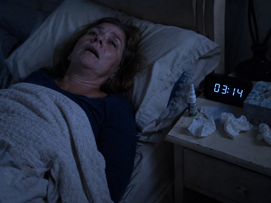 Bildprompt: Fotorealistische Nachtszene — eine Frau um die 55 liegt auf dem Rücken im Bett und atmet mit leicht geöffnetem Mund, angespanntes, unruhiges Gesicht. Auf dem Nachttisch zeigt eine digitale Uhr 03:14, daneben liegen ein Nasenspray und zerknüllte Taschentücher. Schwaches blaues Nachtlicht, dokumentarischer, realistischer Look.