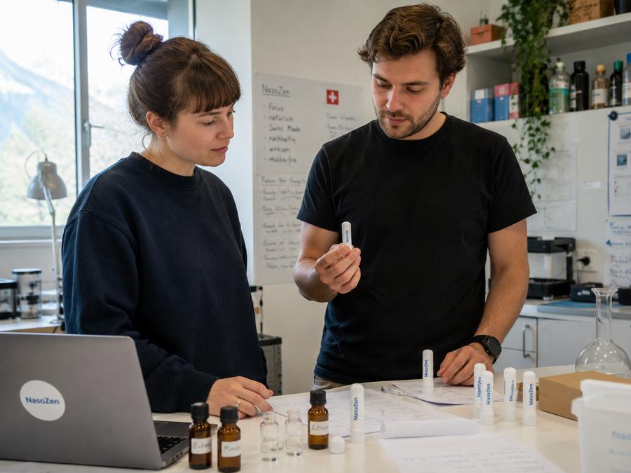 Bildprompt: Fotorealistisches Editorial-Foto im Querformat — ein junger Mann und eine junge Frau Mitte zwanzig (Gründer-Duo, dunkle schlichte Kleidung) stehen in einem kleinen, hellen Schweizer Labor-/Arbeitsraum an einem weissen Tisch mit Notizen, Laptop mit NasoZen-Sticker, braunen ätherischen Ölfläschchen (Etiketten „Eukalyptus", „Minze") und mehreren weissen Inhaler-Stick-Prototypen. Er hält einen weissen NasoZen-Stick (beigefügtes Produktbild als Referenz) prüfend in der Hand, sie schaut konzentriert auf die Notizen. Im Hintergrund ein Whiteboard mit „NasoZen"-Stichpunkten und einer kleinen Schweizer Flagge, Fenster mit Bergblick. Natürliches Tageslicht, dokumentarischer Startup-Reportage-Look, hochauflösend.